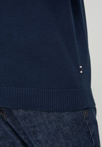 Ciemnoniebieski dziergany dół swetra z prążkowanym brzegiem nad ciemnoniebieskimi dżinsami, z widoczną małą metką materiałową na bocznym szwie swetra.