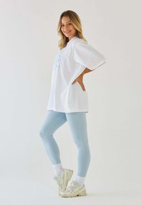 Biała, oversize'owa koszulka z niebieskim wzorem, zestawiona z jasnoniebieskimi legginsami i beżowymi sneakersami. Stylizacja uzupełniona jest o białe skarpetki.