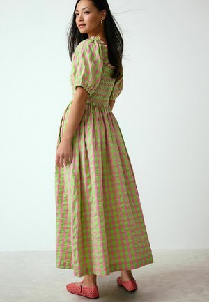 Frau trägt ein grün-rosa kariertes Maxikleid mit Puffärmeln und rosa Cutout-Ballerinas, steht auf einem schlichten hellen Boden und vor einer Wand.