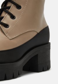 Bottine en cuir beige avec semelle en caoutchouc noir, épais talon carré et fermeture à lacets. Caractéristiques d'un panneau contrastant au niveau du talon et d'une semelle texturée.