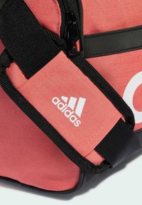 Borsa sportiva in tessuto corallo con dettagli neri, dotata di chiusura con cerniera e logo Adidas impresso in bianco su una tracolla.