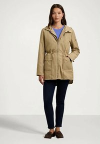 Žena u bež jakni s patentnim zatvaračem i kapuljačom, plavoj majici, tamnim skinny trapericama i smešnim mokasinkama, stoji protiv jednostavne pozadine.