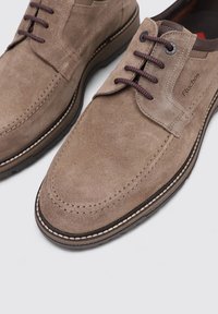 Zapatos de ante gris con punta redondeada, suela oscura contrastante y dos ojales para cordones oscuros. Presentan acentos cosidos y una etiqueta de marca en el costado.