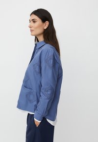 Kobieta z długimi ciemnymi włosami, ubrana w niebieską koszulę zapinaną na guziki z teksturowanym wzorem diamentowym, stojąca w profilu z jedną ręką w kieszeni.