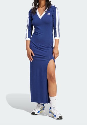 Kobieta ubrana w niebieską sukienkę maxi Adidas z białymi paskami na rękawach i bocznym rozcięciem, zestawiona z białymi skarpetkami Adidas i niebiesko-białymi sneakersami.