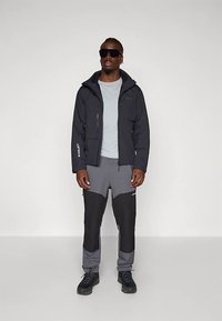 Fekete vízálló dzseki világosszürke ing felett, szürke nadrággal, fekete díszítéssel. A modell napszemüveget és fekete cipőt visel.