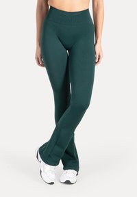 Vrouw die donkergroene leggings met hoge taille en witte sneakers draagt, met gekruiste benen staand tegen een witte achtergrond.