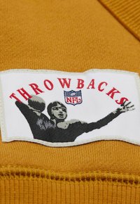 NFL "Throwbacks"-Patch auf einem senfgelben Stoff, mit einem schwarz-weißen Grafik eines Spielers, der einen Football wirft, und rotem Text.
