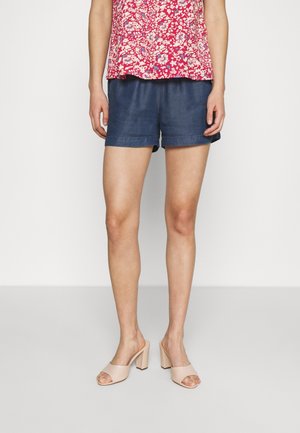 Kvinde iført marineblå shorts, rød blomstret top og beige blokhælede sandaler med åbne tæer, stående mod en ensfarvet lys baggrund.