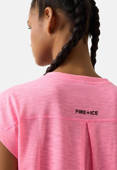 Junge Person mit zwei geflochtenen Zöpfen, die ein rosa Shirt mit dem Text "FIRE + ICE" auf dem oberen Rücken trägt, vor weißem Hintergrund.