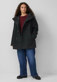 Donkergrijze wollen jas met een brede kraag, dubbele knopen en grote zijzakken, gedragen over een bordeauxrood sweater en blauwe jeans.