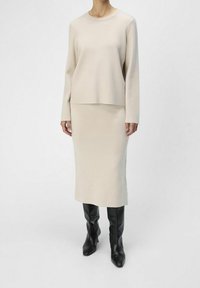 Un haut en tricot beige à manches longues assorti à une jupe midi. Il présente une coupe décontractée et est complété par des bottes en cuir noir bien ajustées.