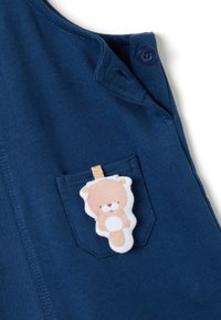 Marineblauer Stoff-Overall mit einer Vordertasche, die mit einem weichen, cartoonhaften Bären-Anhänger verziert ist. Verfügt über einen Knopfverschluss an der Schulter.