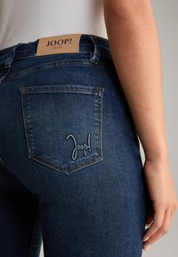 Granatowe dżinsy o średnim stanie, z przeszytym tylnym kieszonką z logo "Joop!" oraz brązową skórzaną naszywką na pasku.