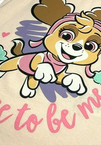 T-shirt présentant un personnage de chien en dessin animé, en tissu beige clair, avec des accents roses et violets, design graphique et texte "Sois moi" en rose.