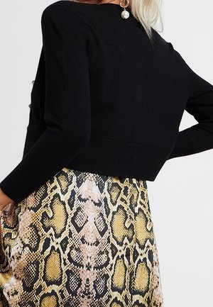 Vrouw die een zwart top met lange mouwen en een hoge taille rok met een geel-zwarte slangensprint draagt, lichtjes zijwaarts gedraaid.