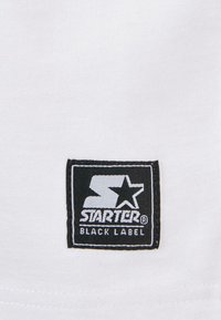 Tissu blanc avec une étiquette tissée noire comportant le logo "STARTER" et le texte "BLACK LABEL" en blanc. Forme carrée, design épuré.