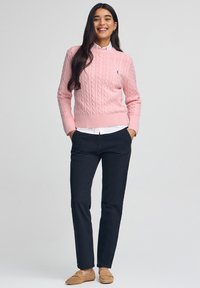 Kvinna som ler, bär en rosa kabelstickad tröja, vit skjorta med krage, svarta byxor och beige loafers, stående med händerna i fickorna.