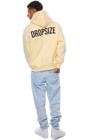 Żółta bluza z kapturem z czarnym napisem "DROPSIZE" na plecach, połączona z jasnoniebieskimi dżinsami i białymi sneakerami. Materiał wydaje się być miękki i ma luźny krój.