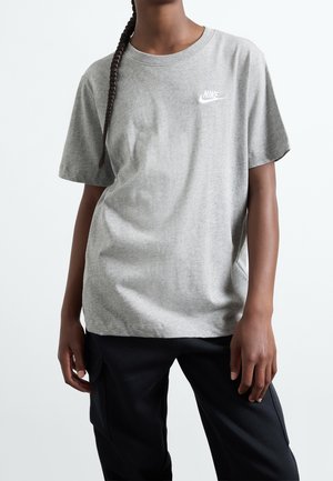 Person trägt ein graues Nike T-Shirt mit Logo auf der Brust und schwarze Hose, steht vor einem schlichten weißen Hintergrund.