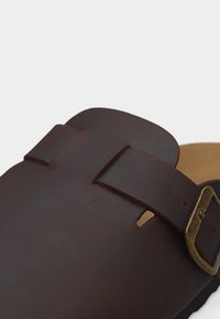 Gros plan sur une lanière de sandale en cuir brun foncé avec une boucle en laiton antique, un lit de pied beige visible en arrière-plan.