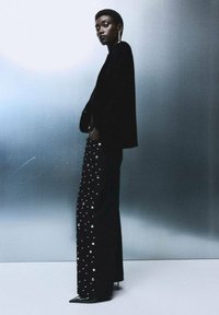 Chaqueta de terciopelo negro combinada con pantalones negros adornados con cristales. Diseño entallado de cintura alta, tacones negros puntiagudos. Textura suave, acentos reflectantes.