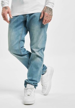 Mand iført falmede blå jeans, hvide sneakers og en hvid langærmet skjorte, med tatoverede hænder synlige mod en ensfarvet baggrund.