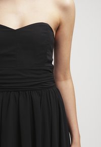 Robe noire sans bretelles avec un corsage ajusté et une taille froncée, portée par une personne sur un fond clair uni.