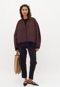 Ruda per didelė striukė su V formos iškirpte, dėvima ant juodo megztinio, derinama su juodomis kelnėmis ir juodomis smailiomis batelėmis. Rankoje geltonas maišelis.