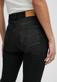 Jeans de mezclilla negra con cintura alta, con una etiqueta de cuero en la parte posterior, costuras mínimas y dos bolsillos traseros. Textura suave y forma ajustada.