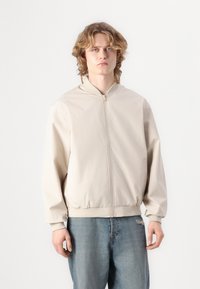 Beige bomberjakke lavet af glat stof, med lynlås foran, ribbede manchetter og bund, båret over lyseblå denimbukser.