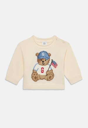 Babypullover in crèmekleur met een beer die een blauwe pet draagt, een wit hemd met een rode "G" en een kleine Amerikaanse vlag vasthoudt.