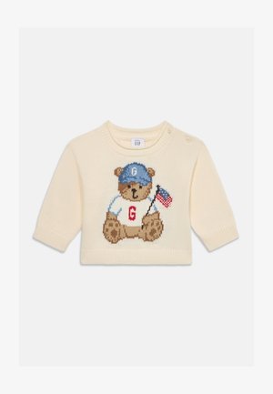 Pull bébé couleur crème avec un ours portant une casquette bleue, une chemise blanche avec un "G" rouge et tenant un petit drapeau américain.
