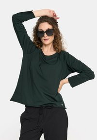 Ciemnozielona bluzka z długim rękawem o luźnym kroju, z dekoltem w serek i subtelnymi detalami. Połączona z czarnymi spodniami.