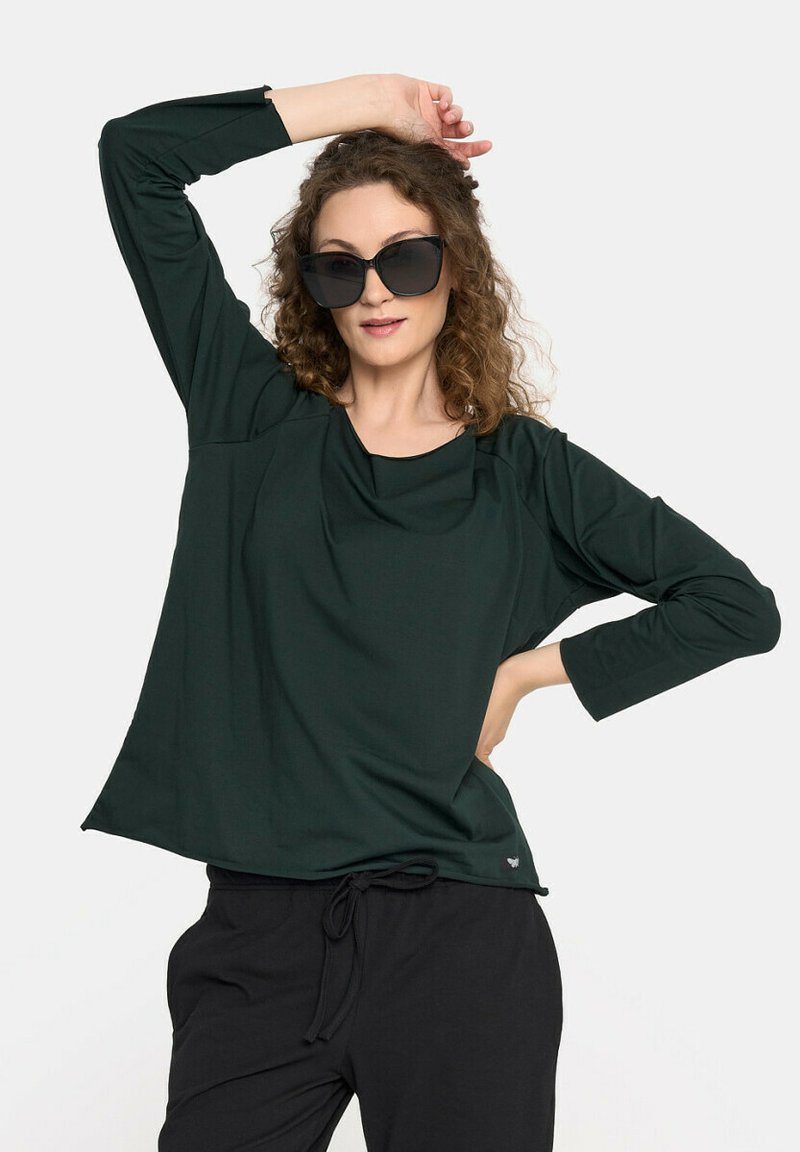 Ciemnozielona bluzka z długim rękawem o luźnym kroju, z dekoltem w serek i subtelnymi detalami. Połączona z czarnymi spodniami.