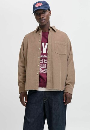 Chemise en velours côtelé marron avec un devant à boutons, une poche poitrine unique, portée sur un sweat à capuche bordeaux avec un texte blanc, associée à un pantalon en denim noir.