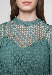 Donna che indossa un top in pizzo verde con un motivo di ritagli floreali e circolari e un collo alto con bordo ondulato.