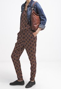 Une combinaison à motifs noir et orange, cintrée à la taille, assortie d'une veste en denim et d'un sac à main en cuir marron. Des baskets noires complètent la tenue.