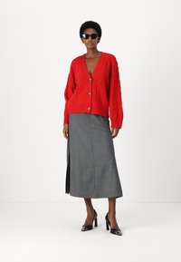 Raudona megzta kardiganas su kabeliais, dideliais mygtukais, derinamas su pilku aukštu įpjovu sijonu. Juodi aukštakulniai bateliai ir saulės akiniai užbaigia išvaizdą.