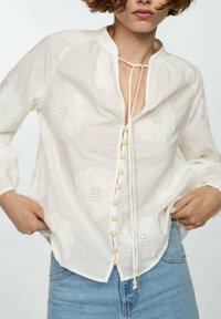 Bílá vyšívaná blůza s květinovými vzory, kulatým výstřihem, stahovací šňůrkou a dřevěnými knoflíky vpředu; lehký materiál.