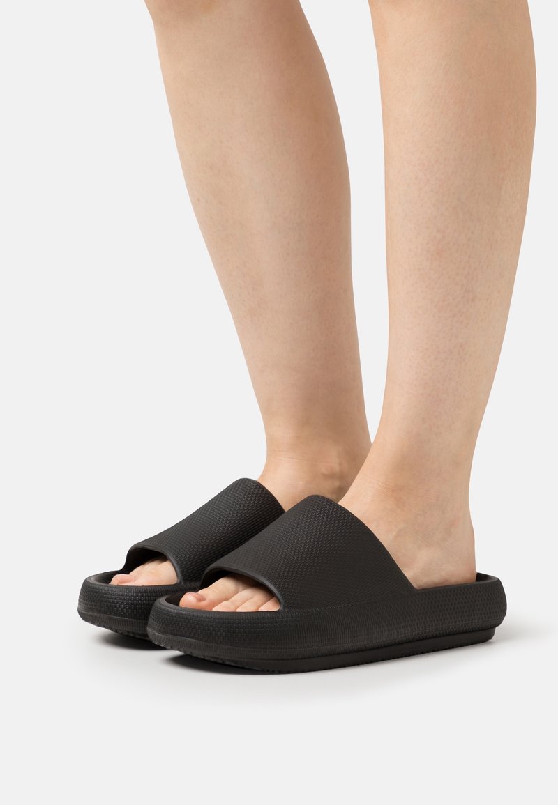 Pieds chaussés de sandales noires à semelles épaisses et ouvertes à l'avant, avec une tige texturée, sur fond blanc.