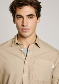 Camicia button-up beige con colletto rilassato e taschino sul petto. Tessuto morbido, vestibilità standard, indossata sopra una camicia azzurro chiaro.