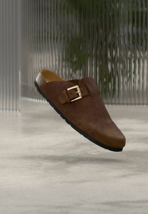 Barna szarvasbőr bebújós cipő arany csattal és fekete talppal, elmosódott, texturált üveg háttér előtt, sima padlón.