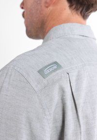 Chemise gris clair avec une texture tissée, arborant une petite étiquette verte Oxbow sur l'épaule et un design de col classique.