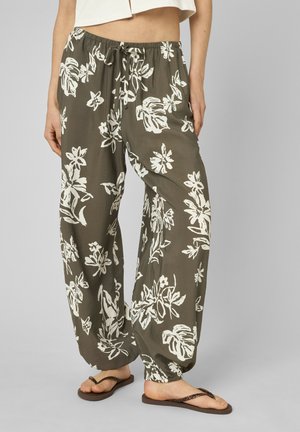 Pantalon ample vert olive avec motifs floraux blancs, taille à cordon, poignets élastiques, porté avec des tongs marron foncé et un crop top blanc.