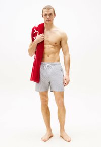 Gestreifte Badeshorts in Blau und Weiß mit elastischem Bund und Kordelzug. Das Modell hält ein rotes Handtuch, hat barfuß und einen neutralen Hintergrund.