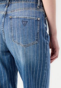 Jean en denim bleu avec des rayures blanches verticales et un patch en forme de triangle sur la poche arrière. Le tissu a une texture lisse.