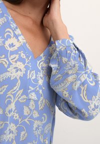 Blouse bleu clair avec un motif floral en crème et or. Dotée d'un décolleté en V, de manches bouffantes et d'un tissu texturé.