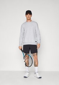Felpa grigia con polsini a coste, berretto nero, pantaloni corti blu e scarpe da ginnastica bianche. Tiene in mano una racchetta da tennis, mostrando tatuaggi sulla parte inferiore della gamba.