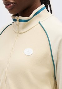 Κοντινή λήψη ενός ατόμου που φοράει μπεζ μπουφάν με φερμουάρ, διακοσμημένο με τυρκουάζ περιθώρια και ρίγες στον γιακά, με ένα μικρό στρογγυλό λευκό λογότυπο στο στήθος.